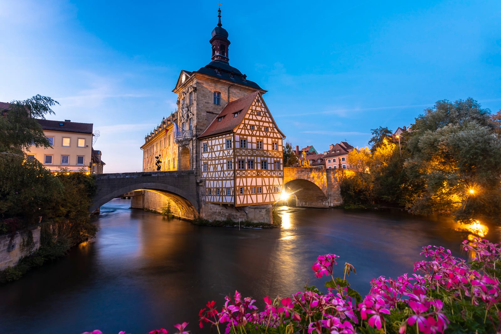Das Alte Rathaus von Bamberg auf der Regnitzinsel, abends beleuchtet, mit Brücke und Blumen im Vordergrund